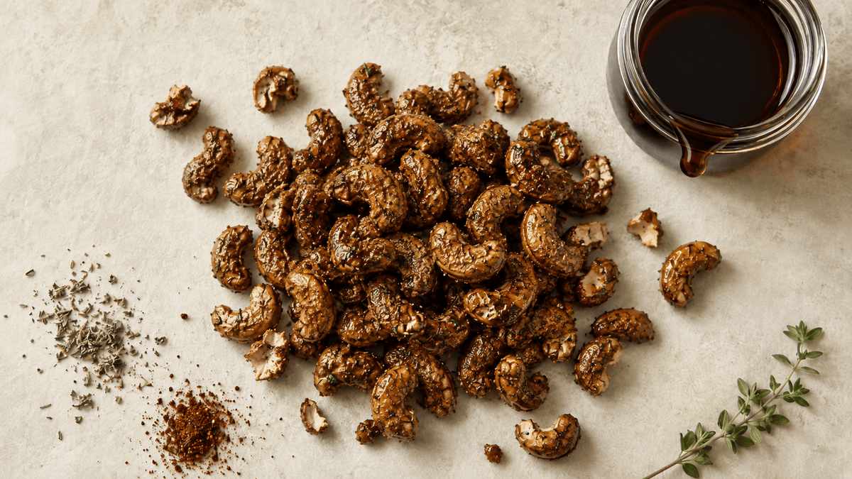 Roasted cashews with a dark set maple coating holding spice flecks, beside a jar of dark maple syrup on a cream background.